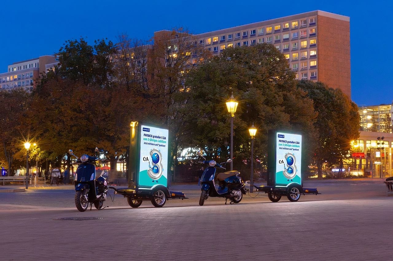 Nachtaufnahme von zwei beleuchteten City-Light-Postern auf Anhängern mit Vespa-Zugfahrzeug Nachtaufnahme von zwei beleuchteten City-Light-Postern auf Anhängern mit Vespa-Zugfahrzeug